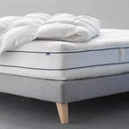 Weiße Matratze Clima Luxe mit feiner Linienstruktur, seitlicher grauer Griff und blauer Biese, liegt auf grauem Bettgestell.