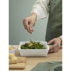 Weiße quadratische ASA Gratinform mit frischem Salat, Person streut Käse-Topping darüber, Holztisch