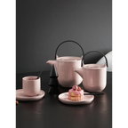 Rosa Teekanne Hanami mit schwarzem Bügel, Tasse und Untertasse sowie kleinem Kuchen auf dunklem Tisch