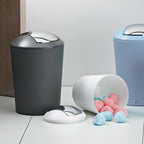 Schwarzer Schwingdeckeleimer mit silbernem Deckel neben weißem umgekipptem Eimer mit rosa und blauen Wattepads auf.
