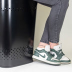 Schwarze Wäschebox Malaga mit Lochdesign, Person sitzt darauf in grauen Jeans und grünen Nike Sneakers