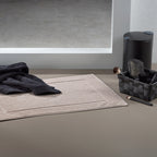 Kela Badvorleger Lavinia Beige Bio auf Badezimmerboden mit schwarzem Handtuch und Accessoires