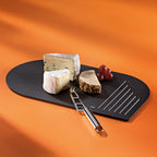 Kela Servier- und Schneidebrett Nobel in Schwarz mit Käse, Trauben und Käsemesser auf orangem Hintergrund