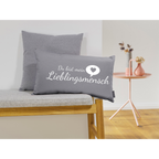 Graues Dekokissen „Du bist mein Lieblingsmensch" auf Holzbank, mit großem Zierkissen und rosé Beistelltisch im Hintergrund.