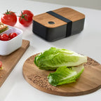 Rundes Holz-Schneidebrett mit Mandala-Gravur, Salat, Lunchbox und Tomaten auf weisser Küchenoberfläche