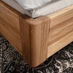 Balkenbett aus Holz mit grauem Cordkopfteil und Metallbeinen auf gemustertem Teppich.