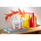 Weiße Elefanten-Dekofigur Baby auf Buch, neben gelbem Vase und bunten Büchern auf Rattantisch