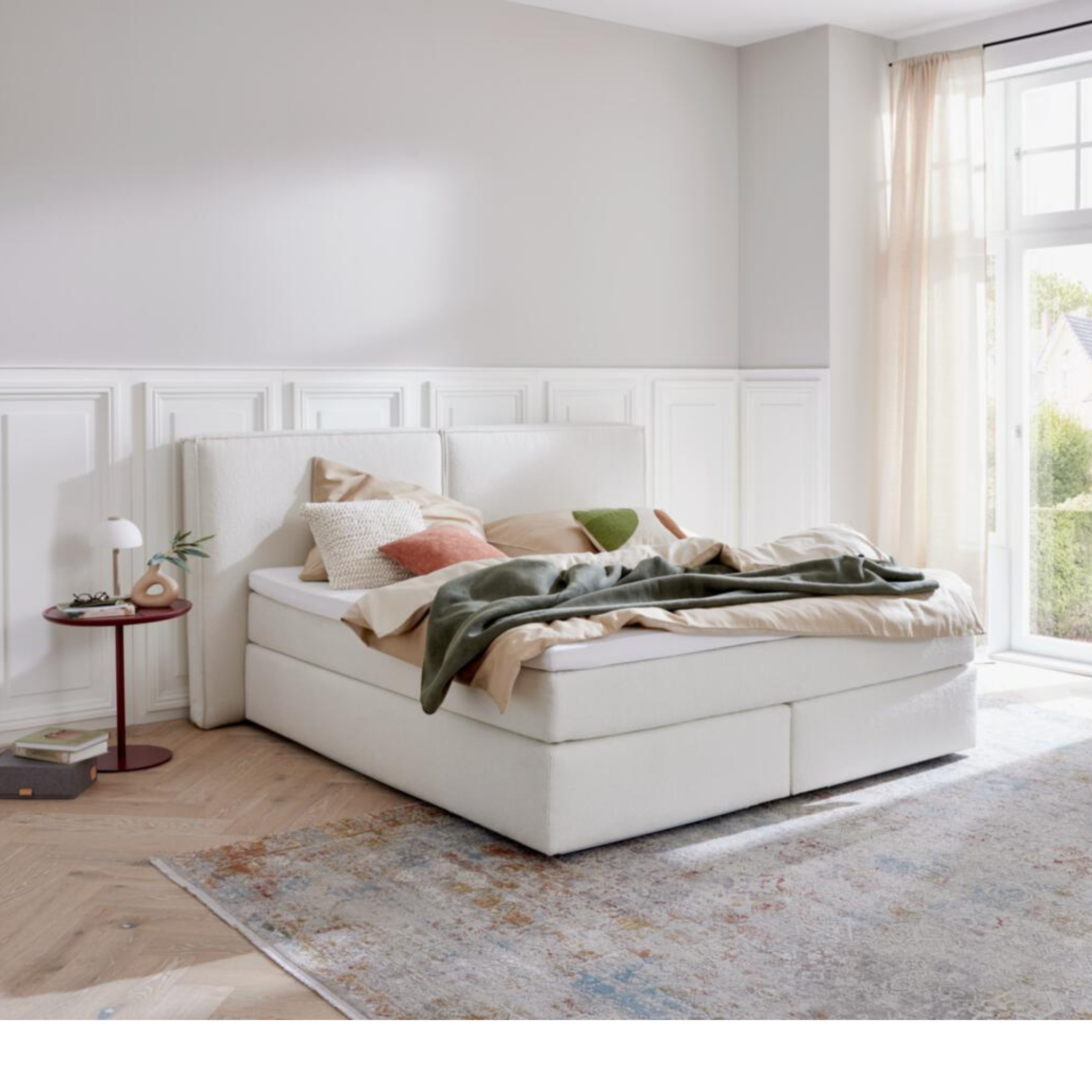 Modernes weißes Boxspringbett 180cm mit hohem Kopfteil, Kissen und Decke in hellem Schlafzimmer mit Tageslicht.