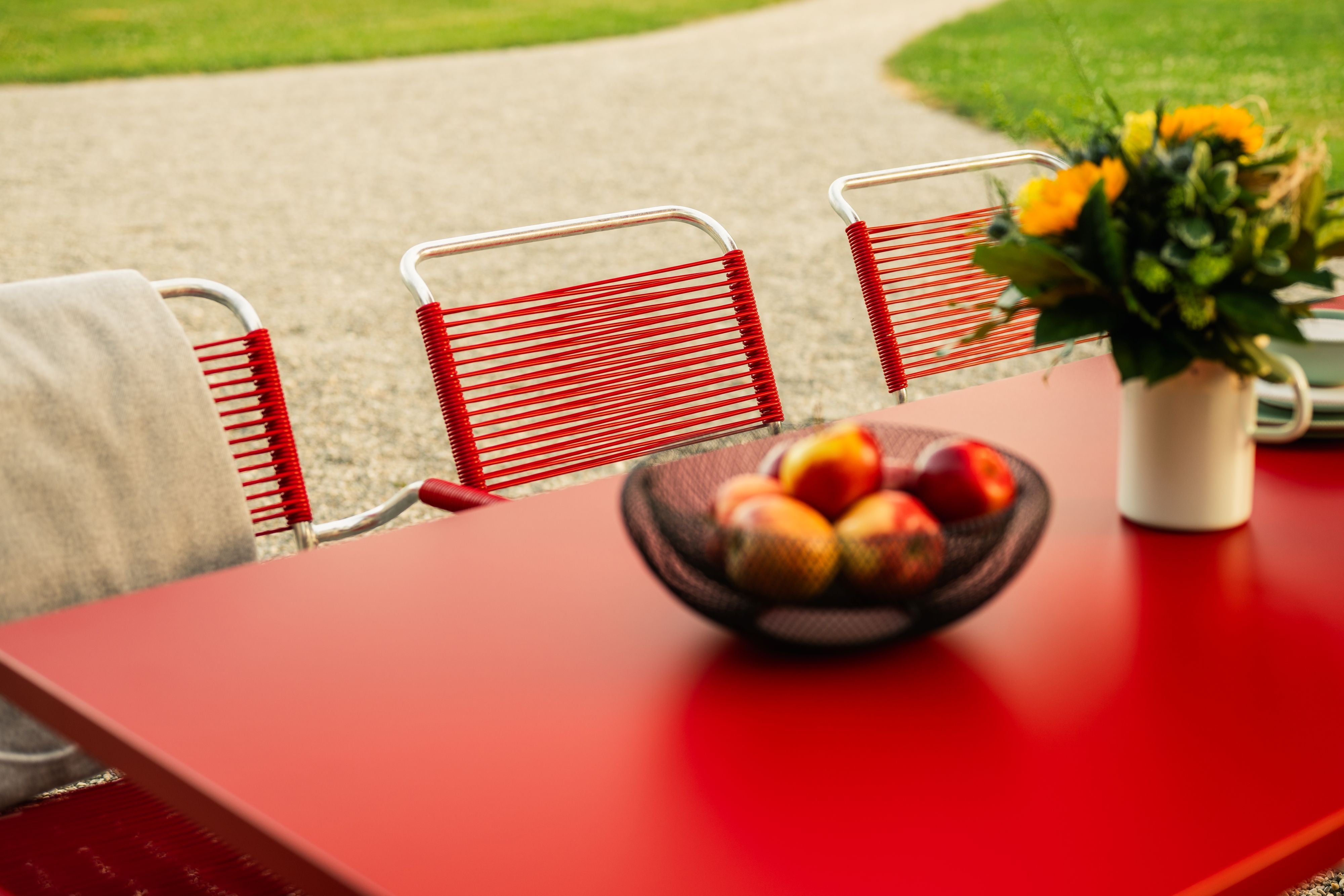 Roter Esstisch mit Spaghetti Stühlen, Obstschale und Blumenvase auf grüner Wiese.