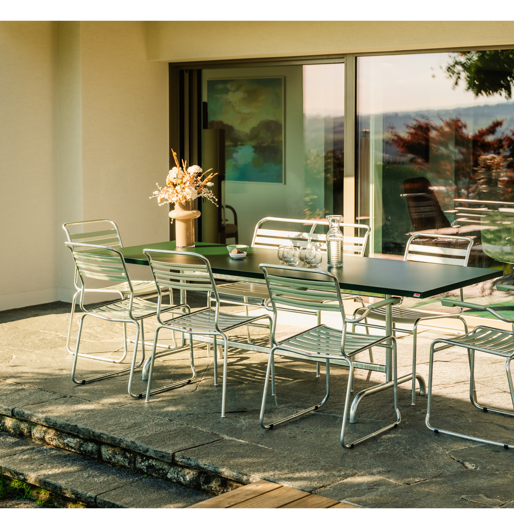 Schaffner Sääntis Lattenstuhl in Pastellgrün auf einer sonnigen Terrasse mit Tisch und großen Fenstern.