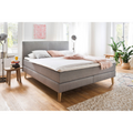 Boxspringbett Greta Porto in Taupe mit Holzbeinen, weisser Bettwäsche und rosa Kissen im hellen Schlafzimmer