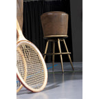 Vintage Barhocker mit braunem Ledersitz und goldenen Metallbeinen neben einem Tennisschläger auf grauem Boden
