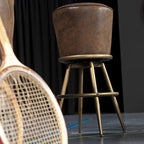 Retro Barhocker mit braunem Lederbezug und goldfarbenem Metallgestell, dekorativ neben einem Tennisschläger